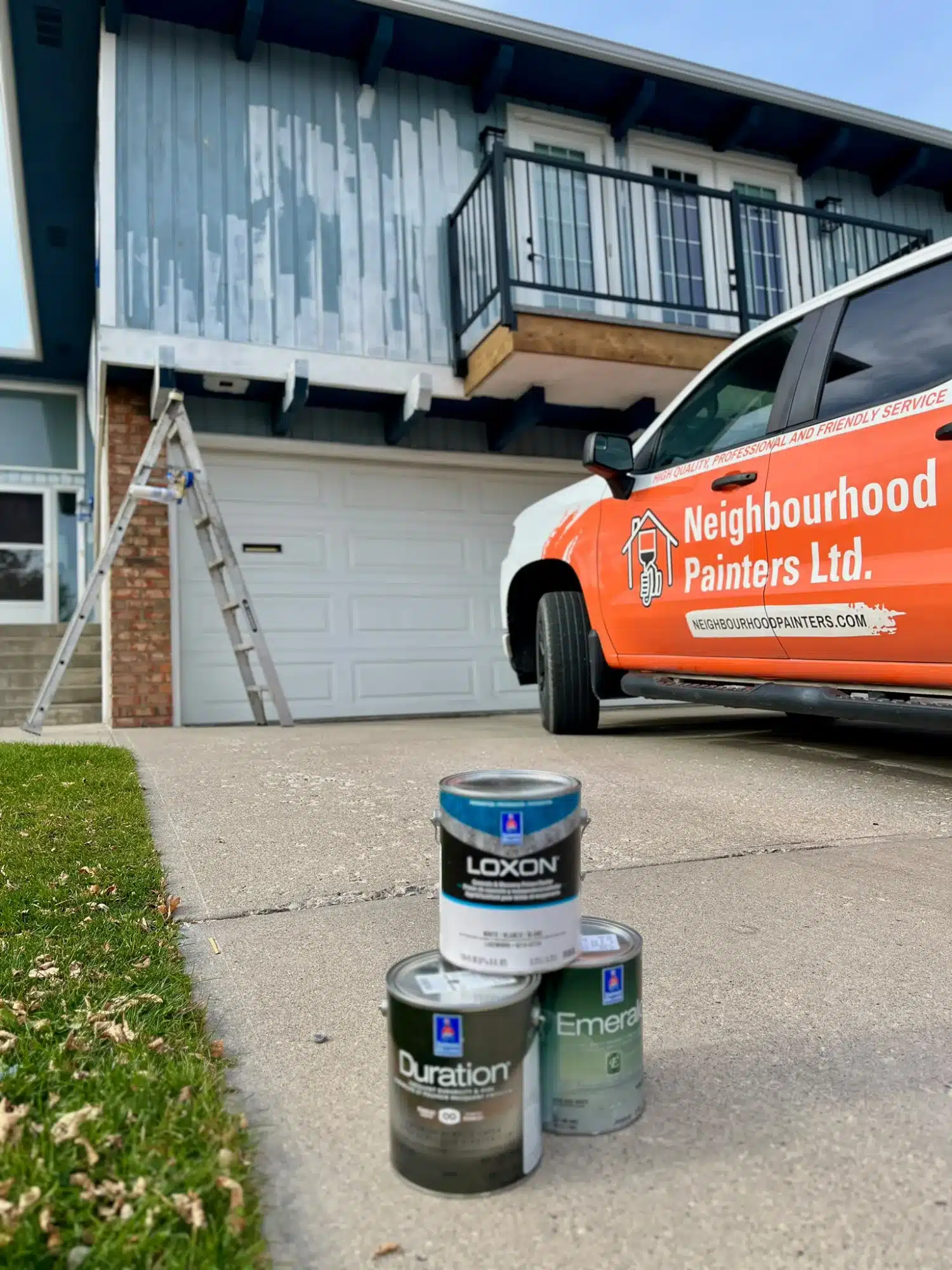 White-painted residential exterior with black metal balcony railing, garage door, and paint cans on driveway in Calgary.